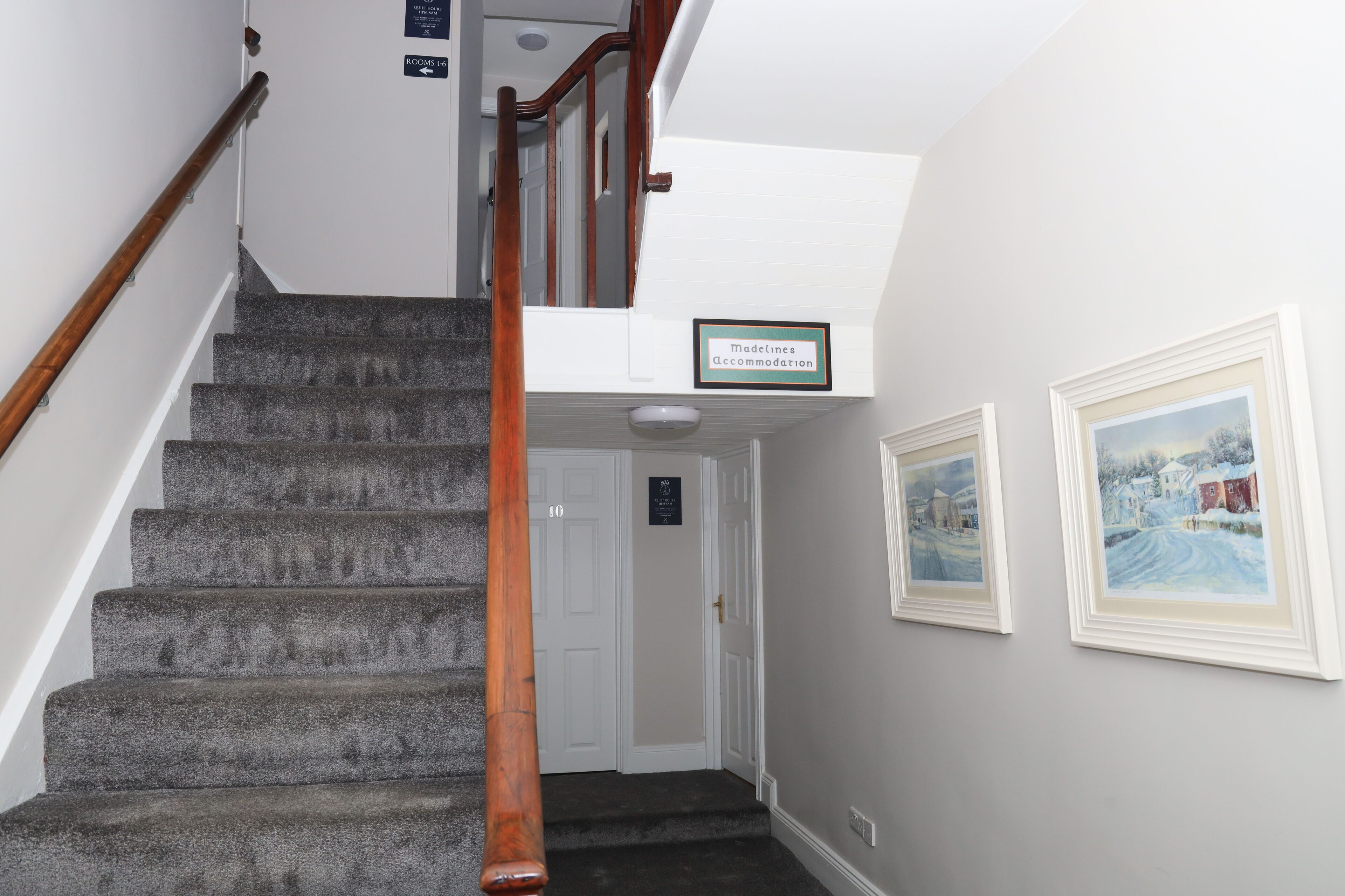 Un escalier avec une rampe en bois mène à une porte portant le numéro « 10 ». À côté de la porte, on trouve une enseigne indiquant « Modeline Accommodation ». Deux tableaux encadrés sont accrochés au mur.