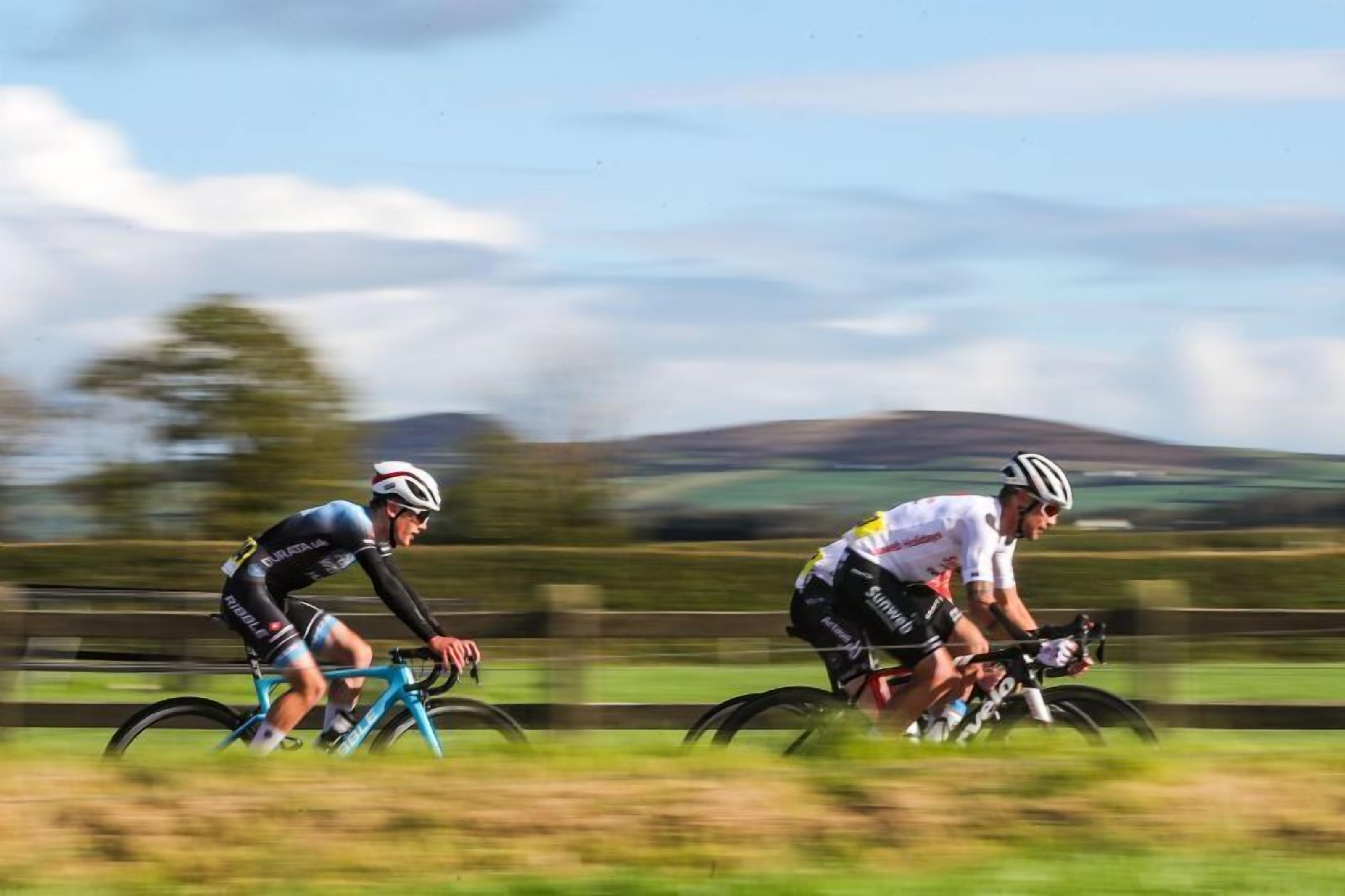 Two cyclists racing on a track with a rural landscape in the background.