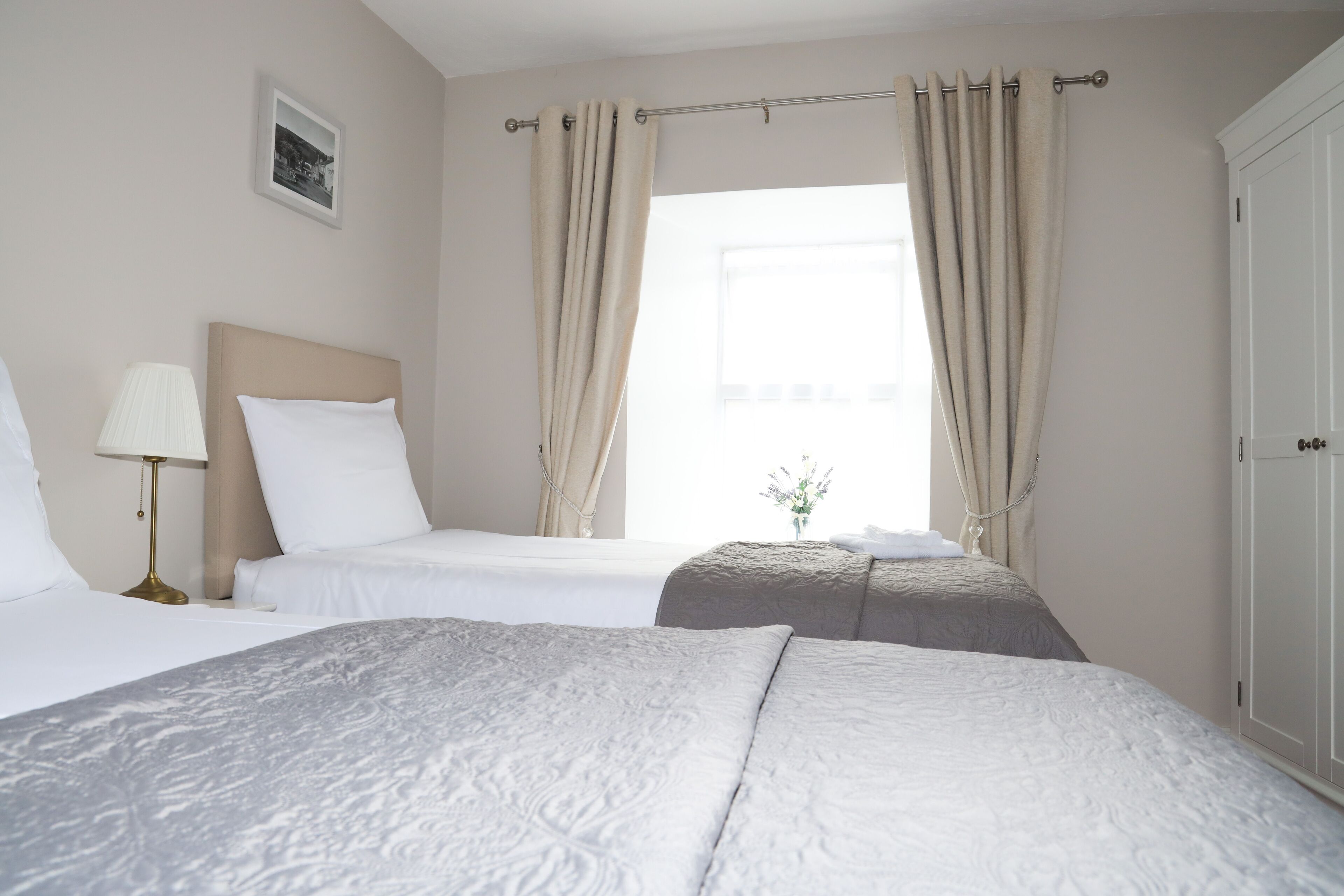 A hotel room with two beds, a bedside lamp, a picture on the wall, and a window with curtains.