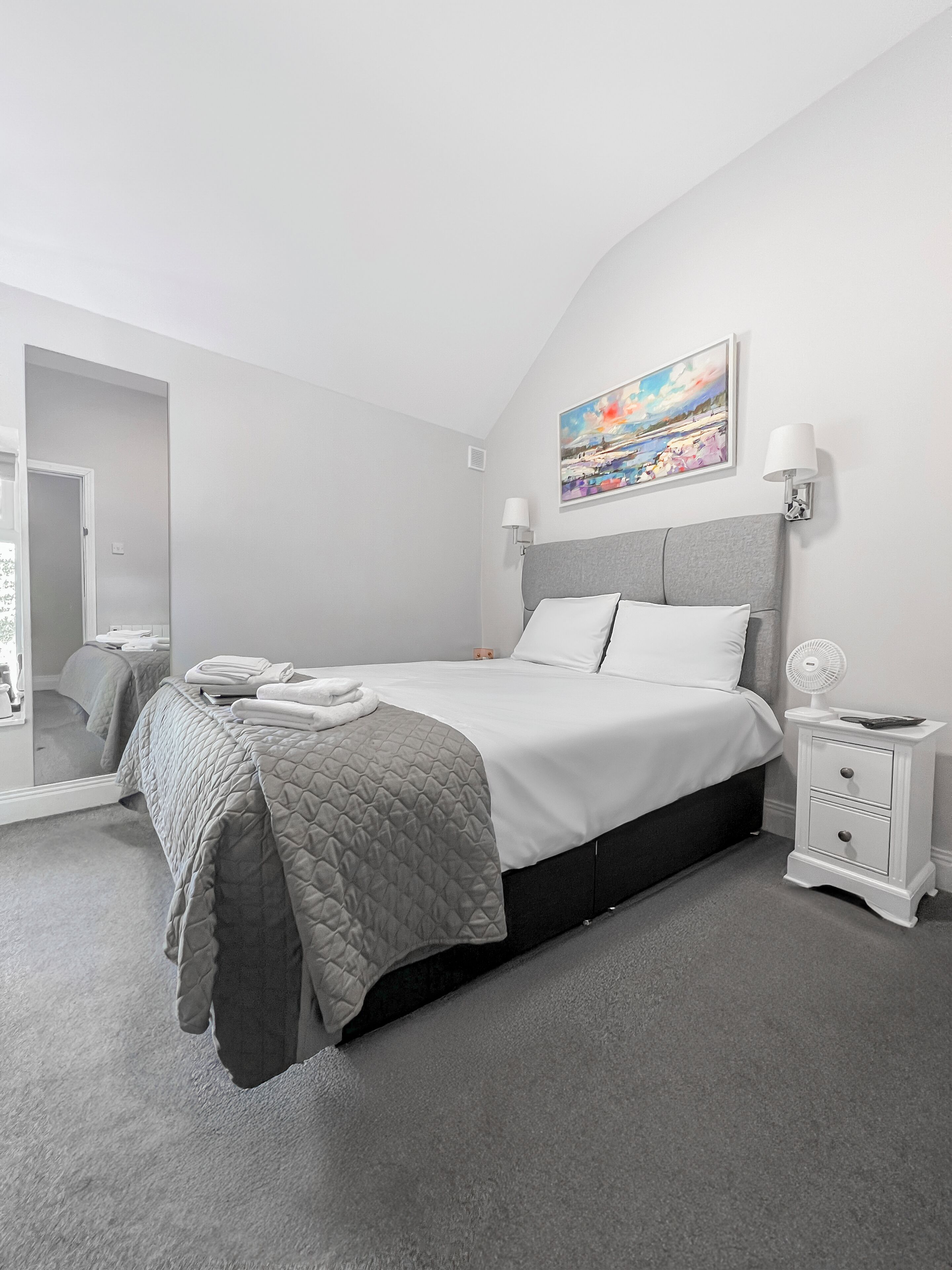 A modern bedroom with a large bed, a bedside table with a lamp, a nightstand, and a painting on the wall.
