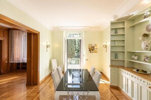 A dining area with a glass-top table and chairs, a view of greenery outside, and a kitchen area with white cabinets and shelves. - Ludovisi Apartment near the Spanish Steps and the Trevi Fountain (Rome)