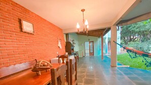 A spacious room with a brick wall, a wooden bench, a dining table, and chairs. There is a chandelier hanging from the ceiling, and a hammock is visible outside the window. - Casa com espaço gourmet, home office e 3 suítes \- Granja Viana (Carapicuiba)