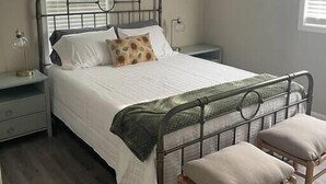 Une chambre à coucher avec un lit en métal, une table de chevet, une chaise, un ventilateur de plafond et deux fenêtres équipées de stores.