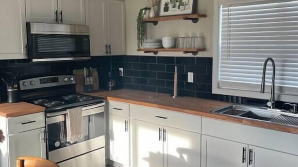 Une cuisine avec des meubles blancs, une cuisinière en acier inoxydable et une fenêtre équipée de stores.