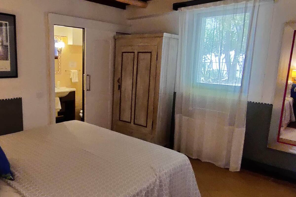Un dormitorio con cama, lámpara de noche, un cuadro enmarcado en la pared, puerta a un baño y ventana con cortinas.