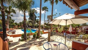 A poolside area with a glass table and chairs, surrounded by palm trees and a swimming pool. - Quinta dos Amigos (Loulé)