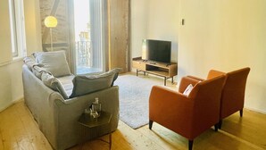 Una sala de estar con un sofá gris, un sillón naranja, un televisor sobre un mueble de madera y un ventanal con vistas a edificios.