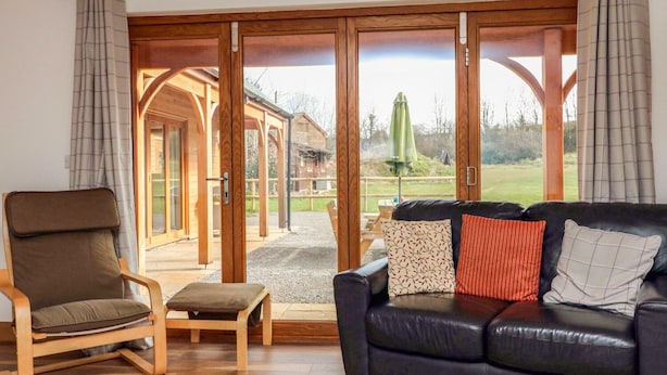 Un salon avec un fauteuil en bois, un canapé en cuir noir et une vue sur un bâtiment en bois, visible à travers de grandes portes vitrées.