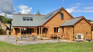 Une maison en bois à deux étages, dotée d’un patio couvert, d’une antenne parabolique et d’une cheminée bien visible d’où s’échappe de la fumée.