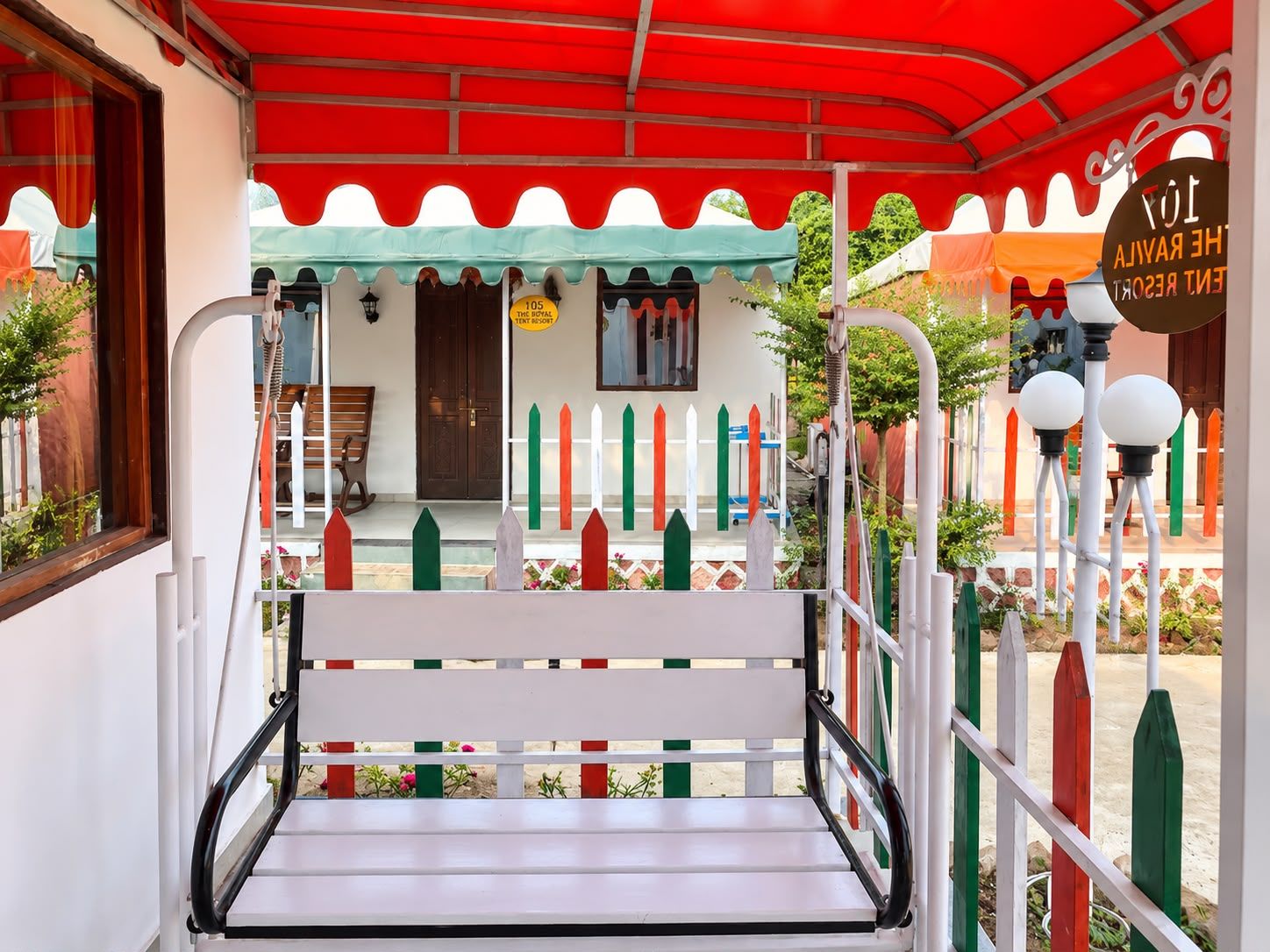 Banco com estrutura metálica e assento branco, posicionado sob toldo vermelho com corrimão branco e postes coloridos.