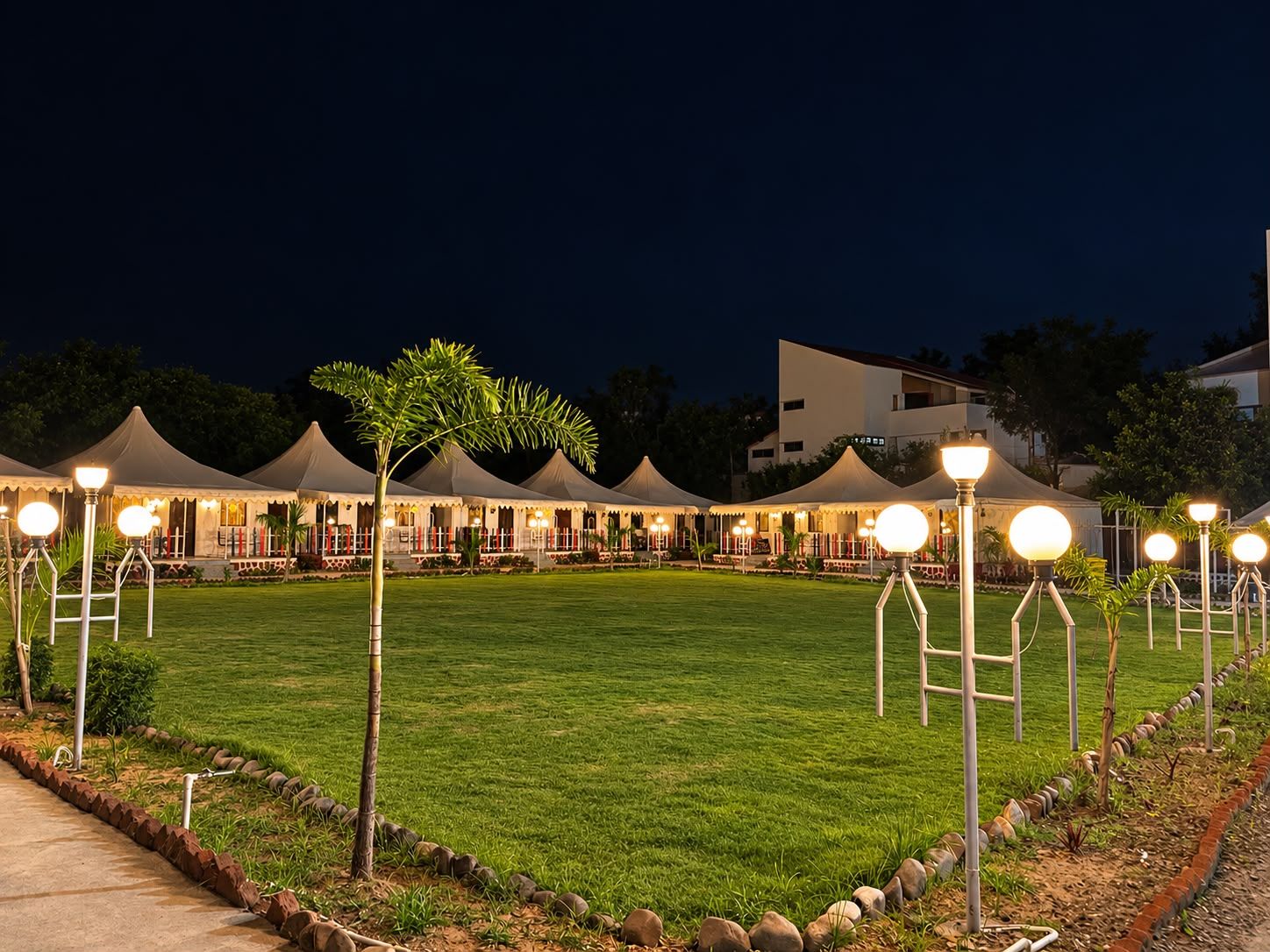 A well-lit outdoor area with multiple tents and a grassy lawn, surrounded by a low stone wall.