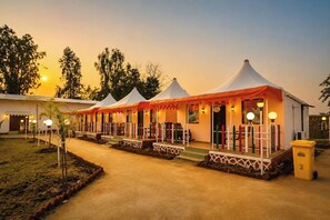 A row of tents with attached verandas, each with a small table and chairs, set against a backdrop of trees and a sunset. - Vasant Vihar Resort (Garudeshwar)