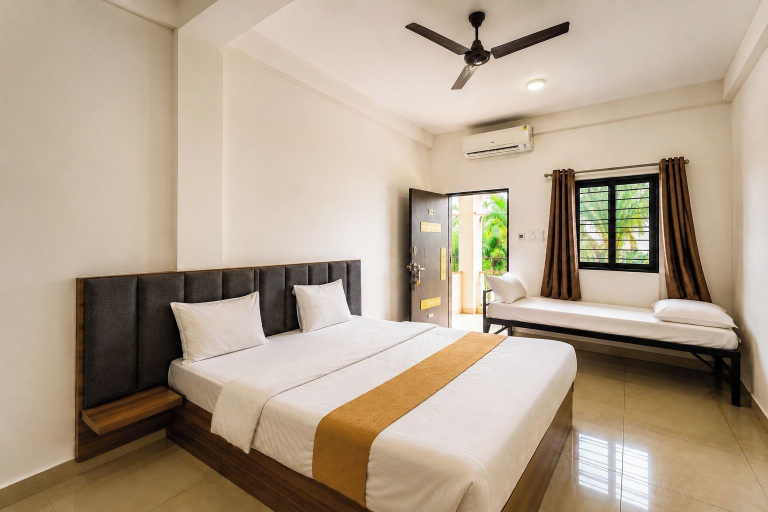 Una habitación de hotel con cama, un banco, ventilador de techo, equipo de aire acondicionado y ventana con cortinas.