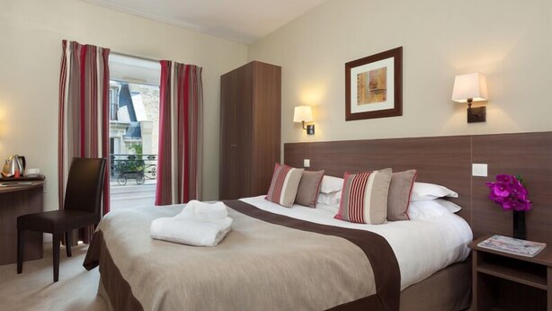 Une chambre d’hôtel avec un lit, un bureau, une chaise, une table de chevet, une lampe, un tableau au mur et une fenêtre avec des rideaux.
