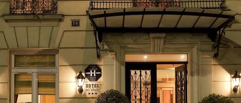 A hotel facade with a dark entrance door, multiple balconies with wrought iron railings, and potted plants.