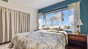 A bedroom with a large bed, a nightstand with a lamp, and a view of palm trees through the window.