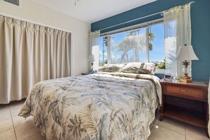 Un dormitorio con una cama grande, una mesita de noche con una lámpara y vistas a palmeras desde la ventana.