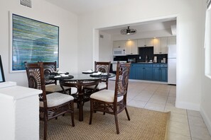 Un comedor con una mesa redonda y seis sillas, una cocina con armarios azules y electrodomésticos blancos, y un cuadro abstracto grande en la pared.