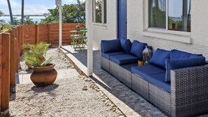 A covered patio with a wicker sofa, a potted plant, and a wooden fence.