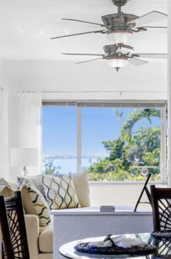 Una habitación con ventilador de techo, un ventanal con vistas al mar, un sofá con cojines estampados y una mesa de comedor con un plato y una taza.