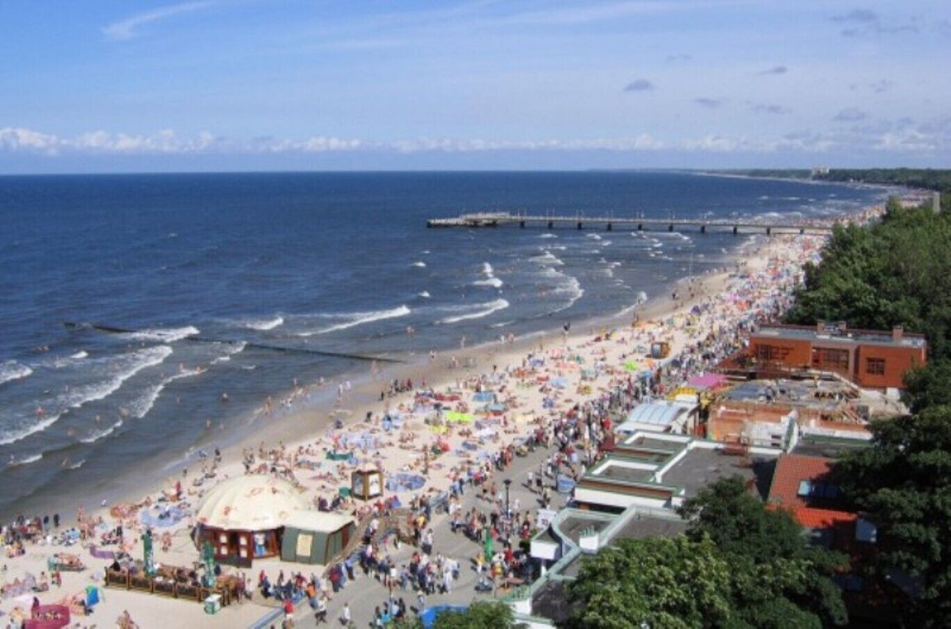 A crowded beach with a pier extending into the sea, surrounded by buildings and greenery.