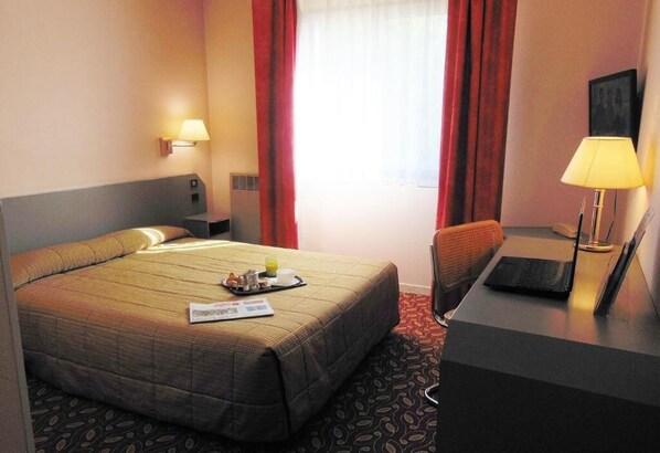 Una habitación de hotel con una cama, un escritorio con una computadora portátil, una silla, una lámpara y una ventana con cortinas.