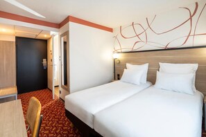 A hotel room with a double bed, a desk with a chair, and a red patterned carpet.