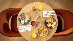 A round wooden table with breakfast items including toast, croissants, and fruit, surrounded by brown leather chairs. - Sure Hotel by Best Western Caen Memorial (Caen)