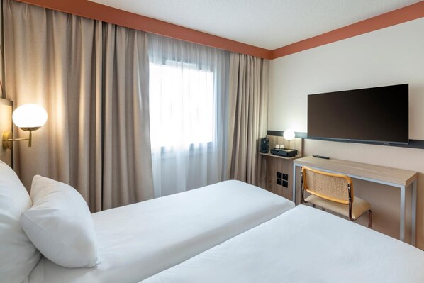 A hotel room with two beds, a desk, a television, and a window with curtains.