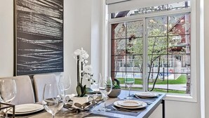Mesa de comedor preparada para seis personas, con platos blancos, copas de vino y cubiertos. Un cuadro abstracto grande adorna la pared, y una ventana muestra vistas a árboles y un edificio.