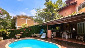 A swimming pool with a wooden deck, surrounded by potted plants and a covered patio area with outdoor furniture. - Casa Alto Padrão na Granja Viana - Cond. Fechado (Carapicuiba)