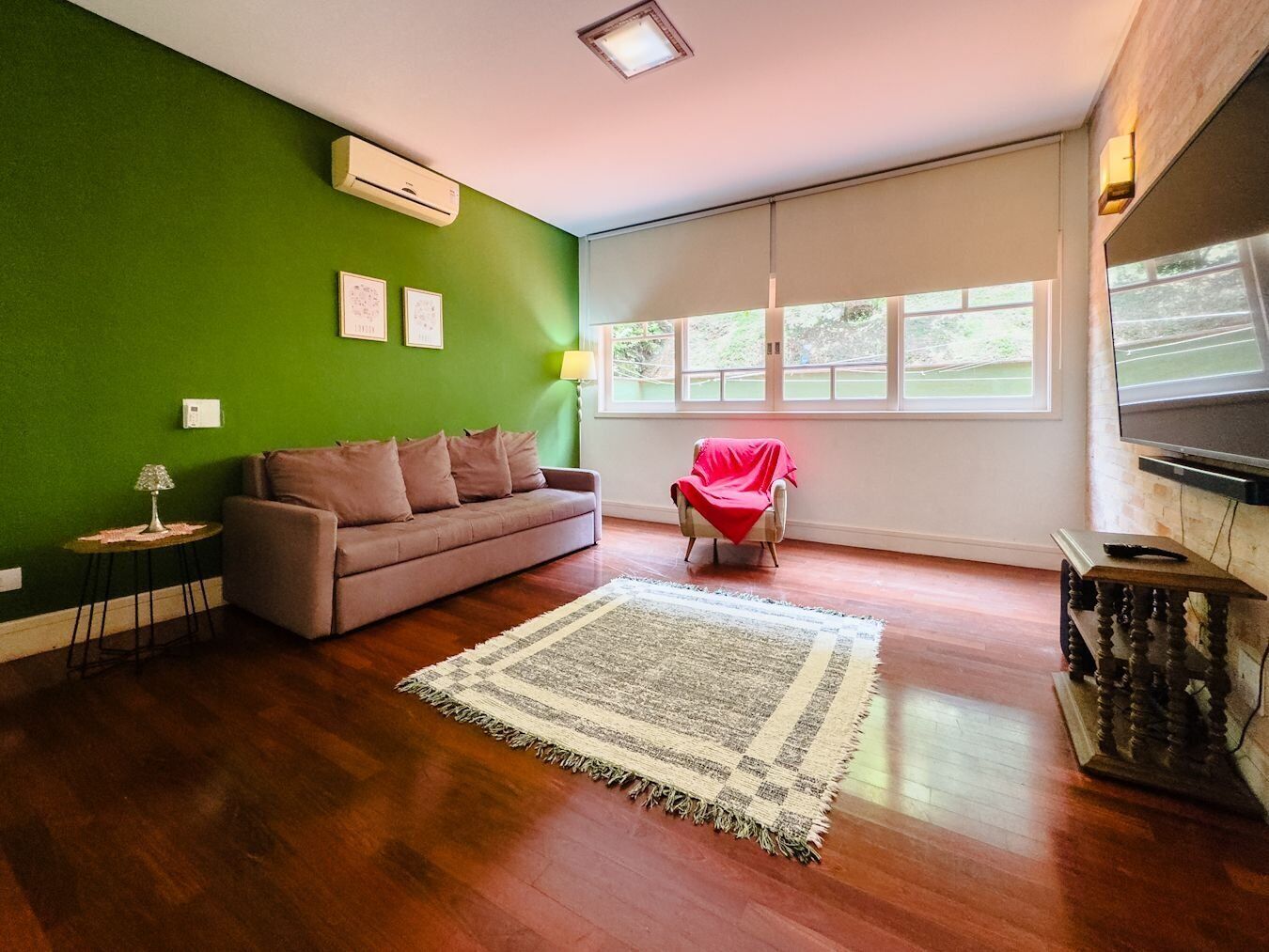 Un salon avec un mur vert, un canapé marron, un fauteuil rouge, une grande fenêtre et un téléviseur à écran plat.