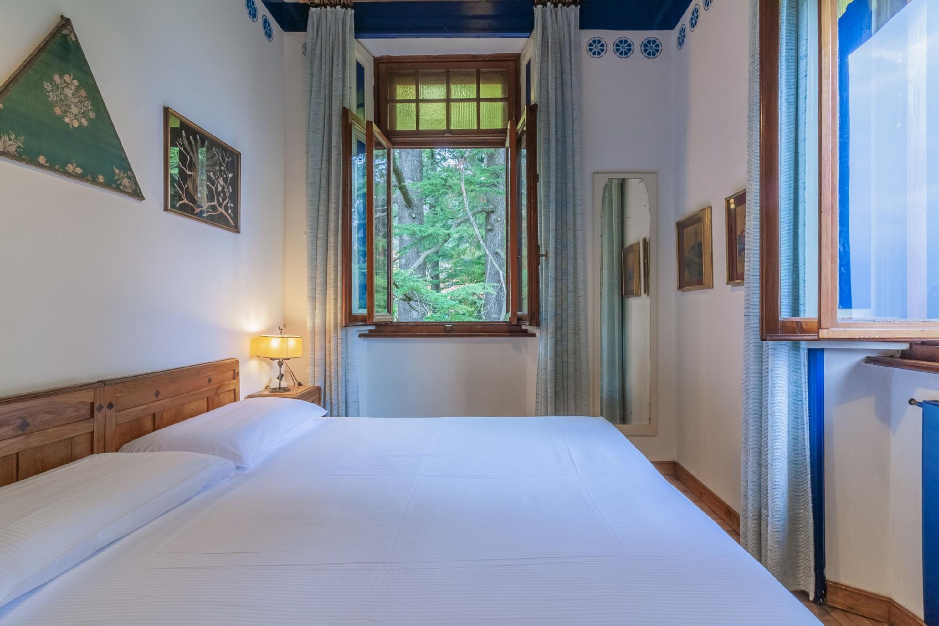 A bedroom with a large bed, a window with curtains, a bedside lamp, and framed pictures on the wall.