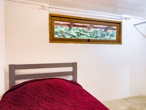 Una cama con colcha roja, cabecera de madera y ventana con vista a follaje.