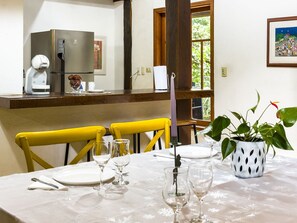Un comedor con una mesa dispuesta para dos personas, un refrigerador y una ventana con vista a follaje.