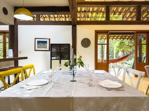 Un comedor con una mesa dispuesta para cuatro, sillas blancas, un aparador con cristalería y una hamaca afuera.