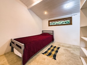 Une petite chambre avec un lit simple, une couvre-lit rouge, une tête de lit en bois, une fenêtre donnant sur un paysage verdoyant et un tapis beige.