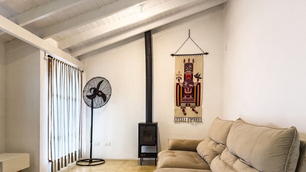 Un salon chaleureux avec un canapé beige, un ventilateur sur pied, un tapis rouge et gris, un plancher en bois et une tapisserie accrochée au mur.