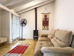 Una sala de estar acogedora con un sofá beige, un ventilador de pie, un tapete rojo y gris, suelo de madera y un tapiz colgado en la pared.