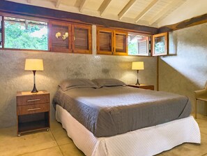 A bedroom with a bed, two bedside tables with lamps, a wooden headboard, and a view of trees through the windows. - Casa com Piscina Privativa e Natureza – Granja Viana, São Paulo (Carapicuiba)