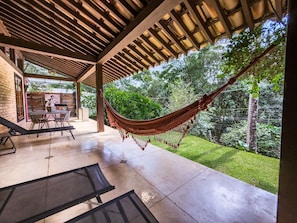 Um pátio externo com uma rede, uma mesa com cadeiras e vista para um jardim exuberante.