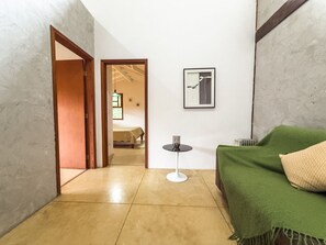 Une petite pièce bien éclairée, avec un lit, un plaid vert, un tableau encadré au mur et une petite table avec une lampe.