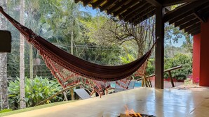 Un hamac sous une pergola en bois, un foyer avec du bois qui crépite, et une terrasse en béton avec de la verdure en arrière-plan.