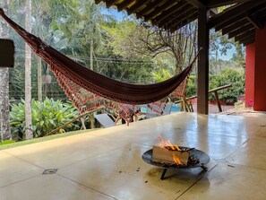 Una hamaca bajo una pérgola de madera, un brasero con leña encendida y un patio de concreto con follaje al fondo.