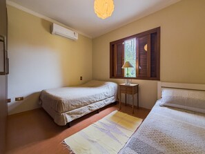 Quarto pequeno e simples com duas camas individuais, uma mesa pequena com abajur e uma janela com persianas.