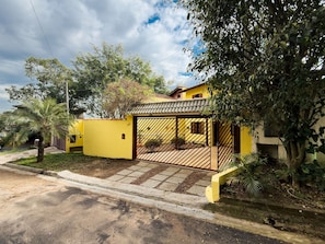 Una casa amarilla con entrada protegida por una reja, rodeada de vegetación y un camino de acceso pavimentado.