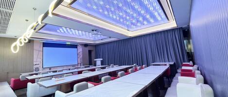 A modern conference room with a large screen, rows of tables, and comfortable seating.