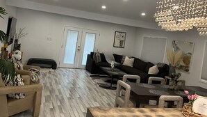 Un salon spacieux agrémenté d’un lustre moderne, d’un canapé noir et d’un coin repas avec une table en bois.
