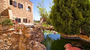 Un bâtiment en pierre doté de fenêtres en arc de cercle et d’une cour intérieure avec un bassin, le tout entouré de verdure et d’un grand arbre.
