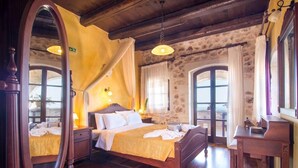 Une chambre à coucher avec un lit en bois, une table de chevet, une commode et un grand miroir. Il y a deux fenêtres avec des rideaux, et on aperçoit un balcon à travers l’une d’elles.